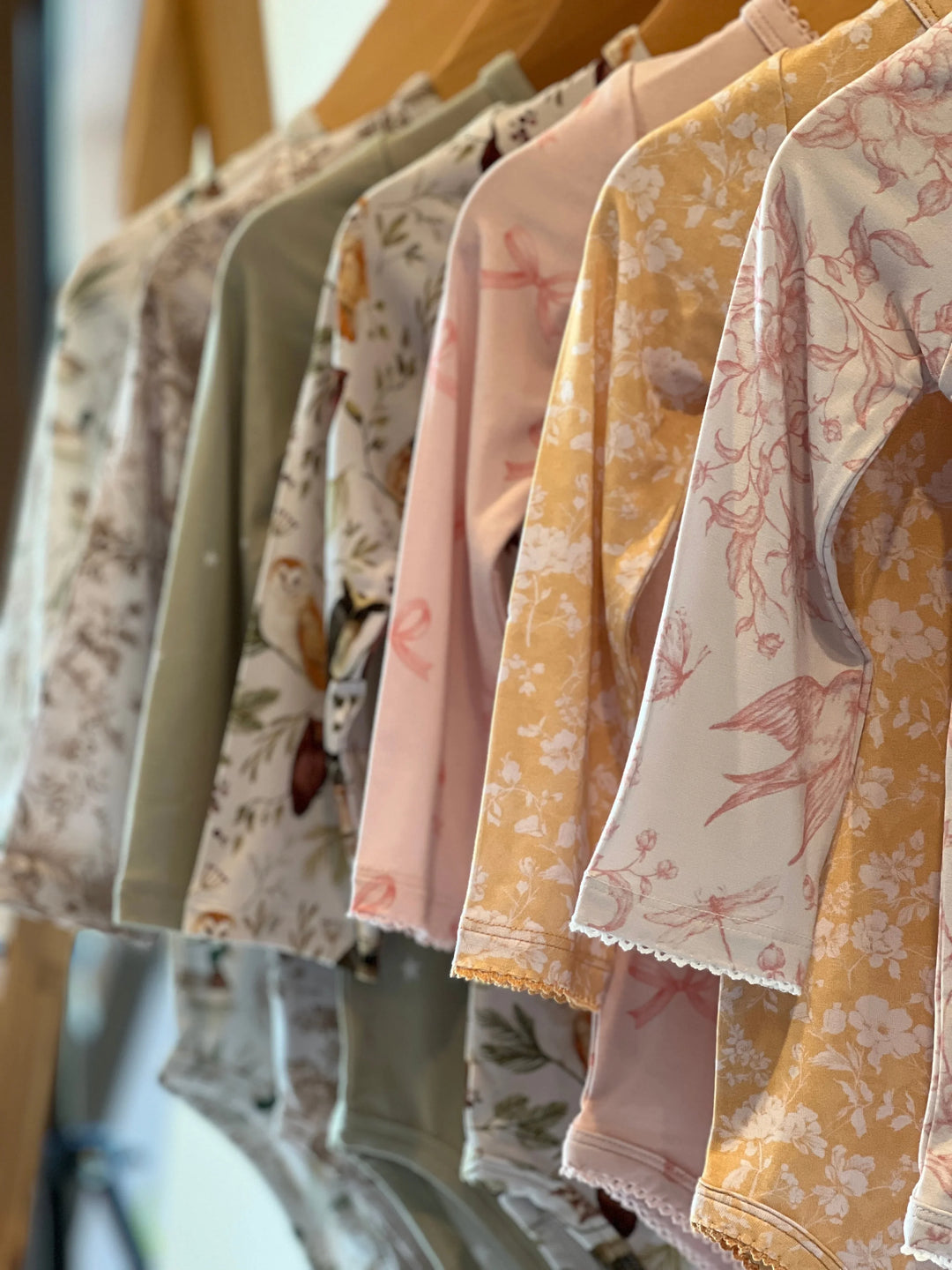 A row of bamboo baby bodysuits in seven nursery-inspired prints from Autumn Moon Kids Clothing, including Woodland Toile, Sage Stars, Botanical Lullaby, Golden Afternoon, Pink Bows, Sleepy Hollow Owls, and Mallard Ducks.