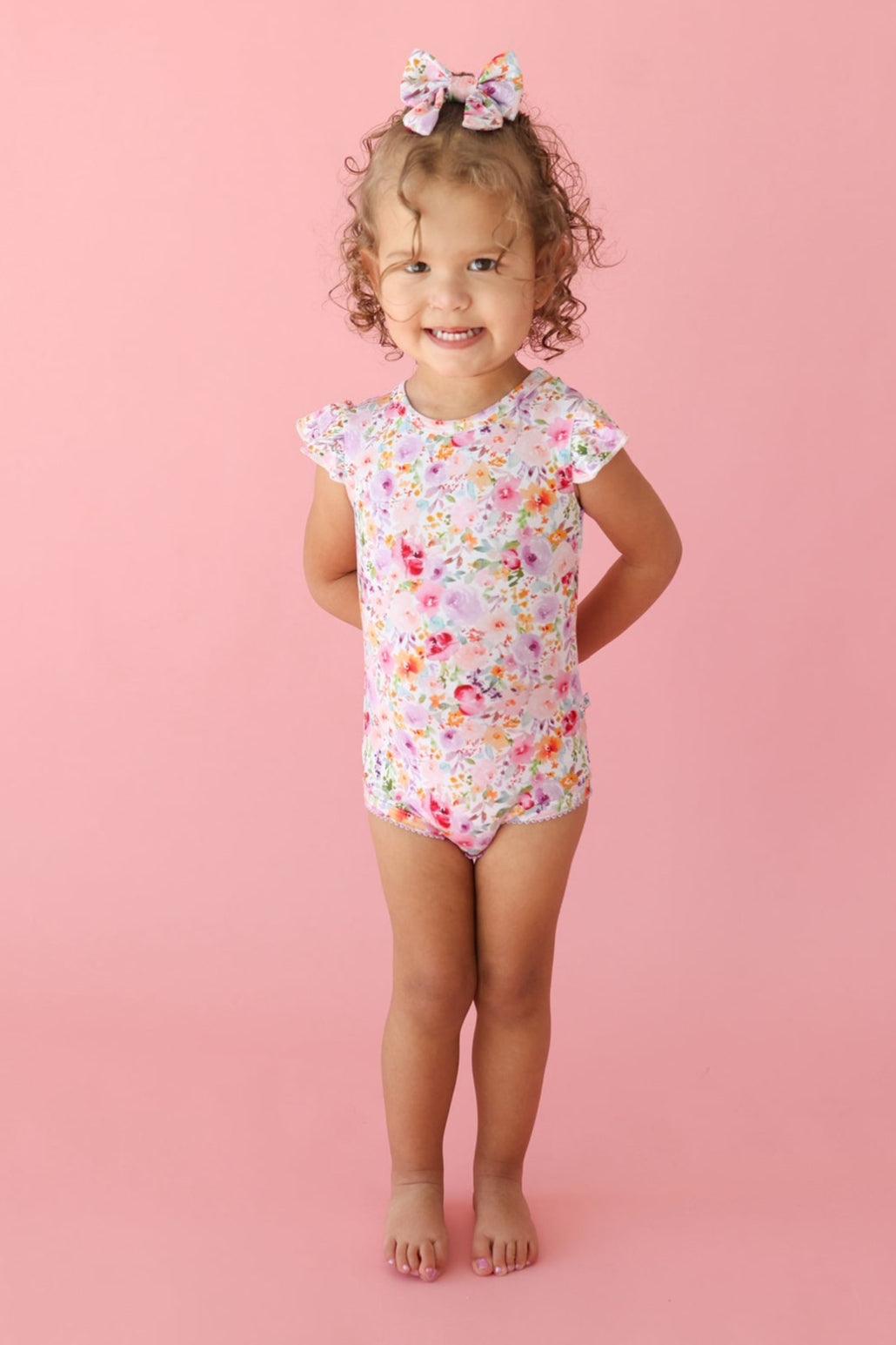 Toddler girl standing in flower garden bamboo bodysuit with ruffle sleeves and floral print, shown in playful pose.