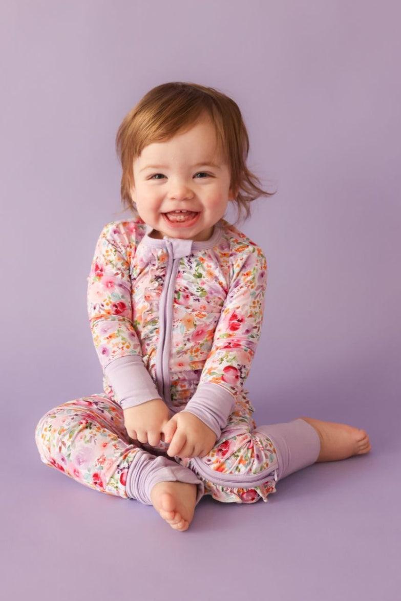 Baby girl sitting in flower garden bamboo zipper pajamas with long sleeves, dual zipper, ruffle butt, and fold-over convertible feet.