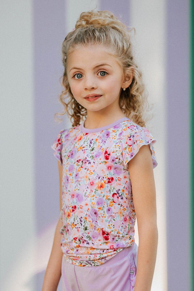 Girl wearing flower garden bamboo T-shirt with short ruffle sleeves, front-facing view in pink floral print.