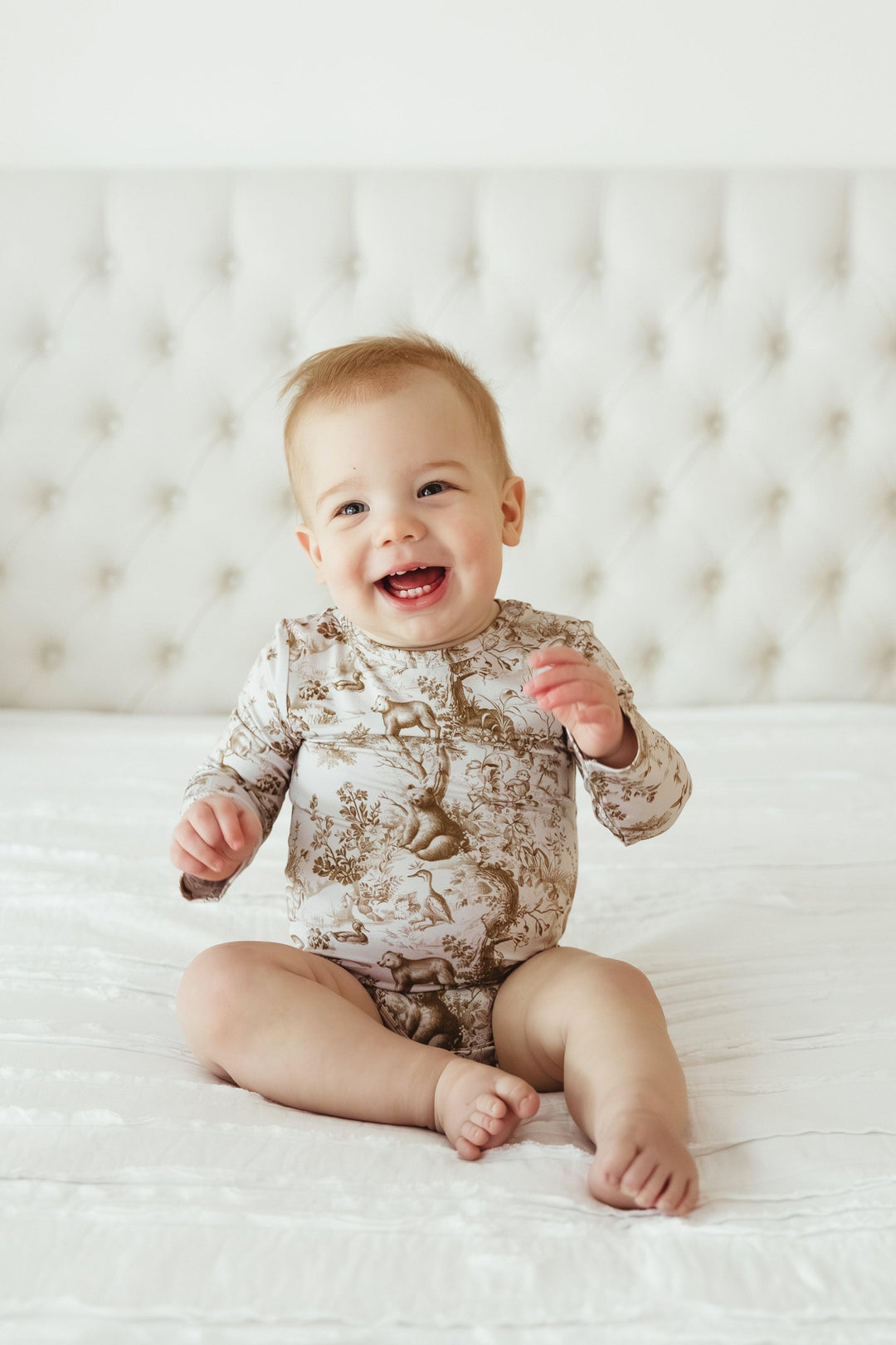 Smiling baby boy wearing Woodland Toile long sleeve bamboo bodysuit, sitting on a white bed — soft and stretchy unisex one-piece ideal for everyday comfort.