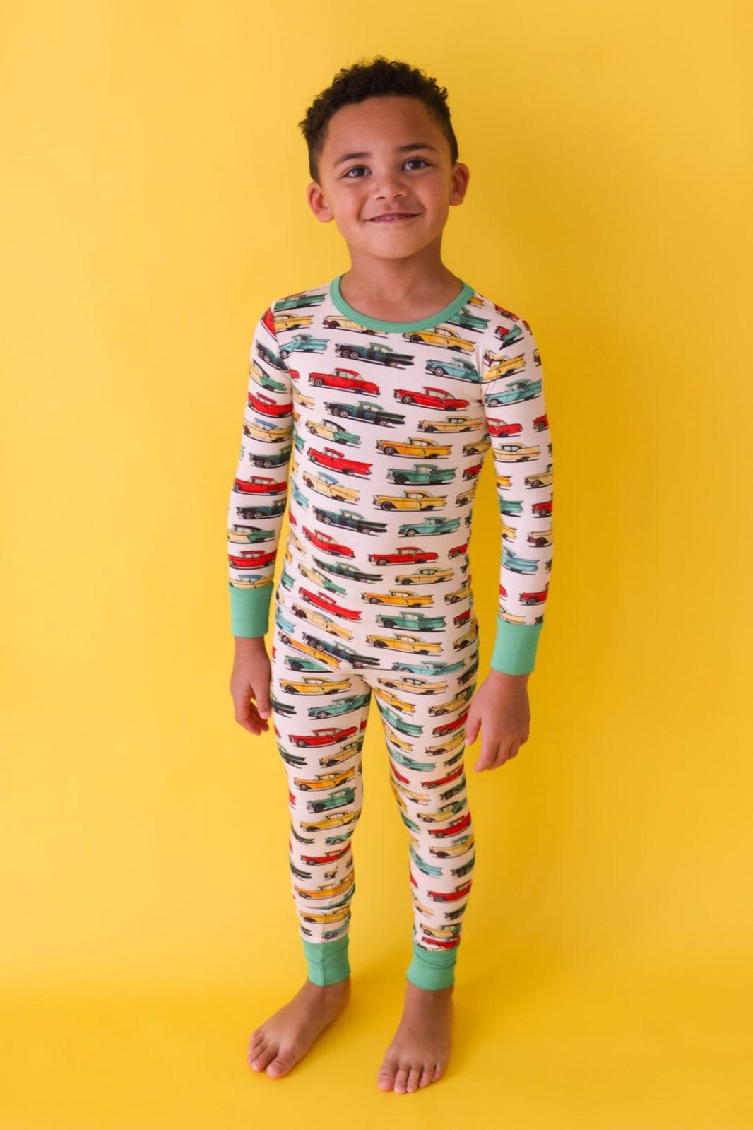 Front-facing photo of little boy in classic cars bamboo pajama top and pants, smiling and standing in soft, stretchy sleepwear with retro car design.