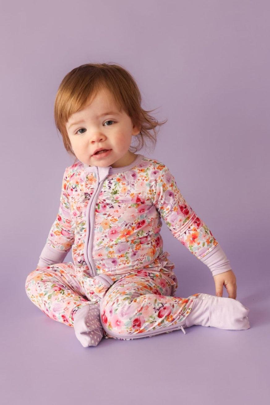Seated toddler in flower garden print bamboo footie pajamas with convertible feet and ruffle bottom design.