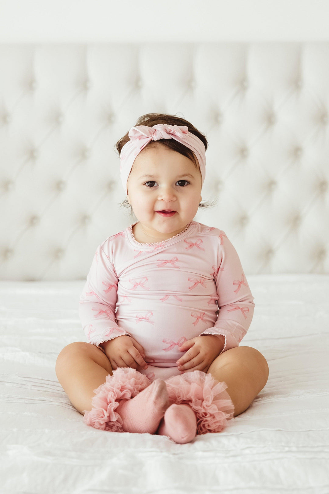 Toddler girl sitting on a bed in pink bows long sleeve bamboo bodysuit and matching headband — cozy, stylish, and perfect for everyday comfort or photos.