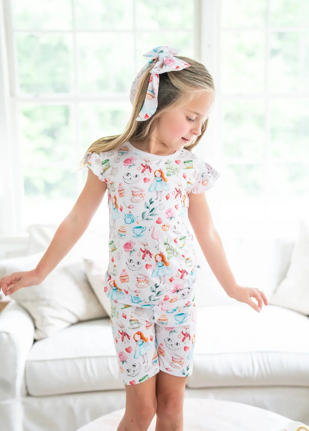 Girl modeling Alice in Wonderland-inspired two-piece pajamas with teacup and cupcake print, shown standing front-facing.