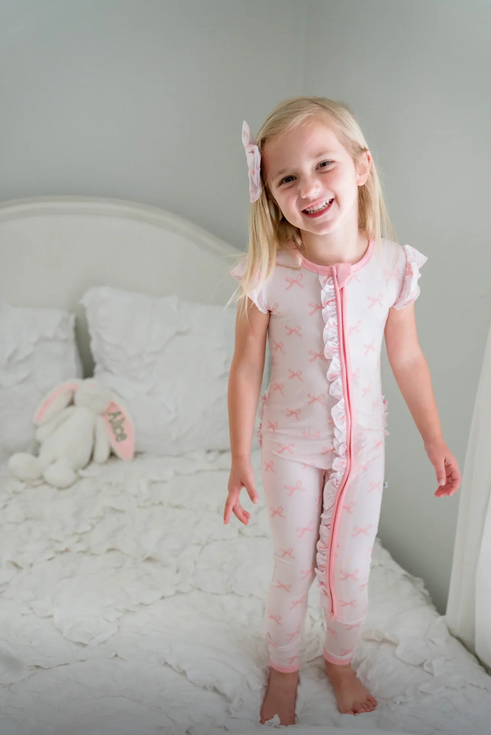 Front view of toddler in pink bows short ruffle sleeve bamboo romper with dual zipper and fold-over feet.