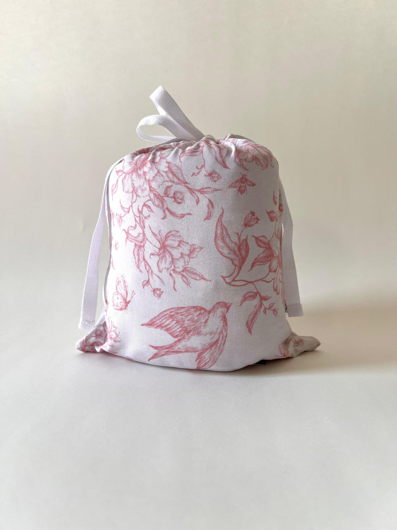 Botanical Lullaby bamboo crib sheet folded inside matching drawstring pouch, featuring a vintage-inspired rose and off-white toile print with birds, flowers, bees, and hummingbirds — gift-ready and perfect for baby registries.
