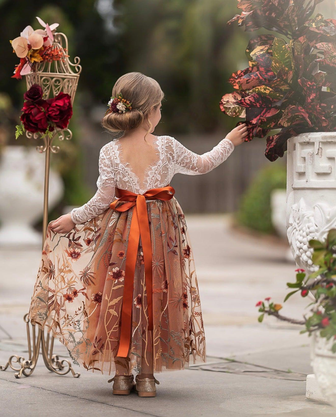 Woman in a floral dress with an orange ribbon standing next to decorative floral arrangements.