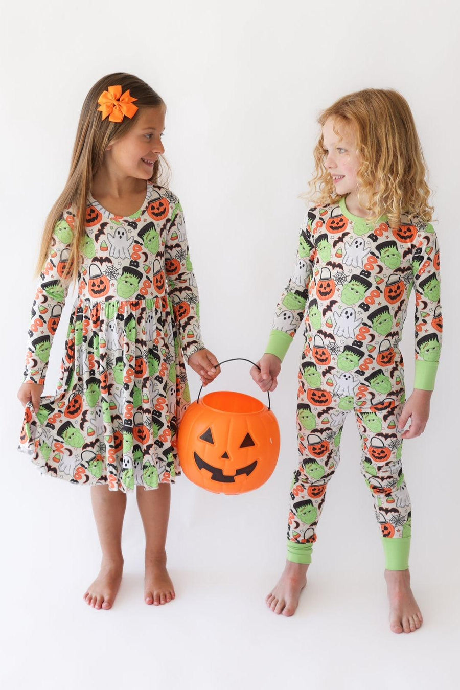 Two young girls standing barefoot against a plain white studio background, wearing matching Halloween-themed long-sleeve pajamas with a print of pumpkins, ghosts, and green monsters. One girl holds an orange jack-o'-lantern bucket.