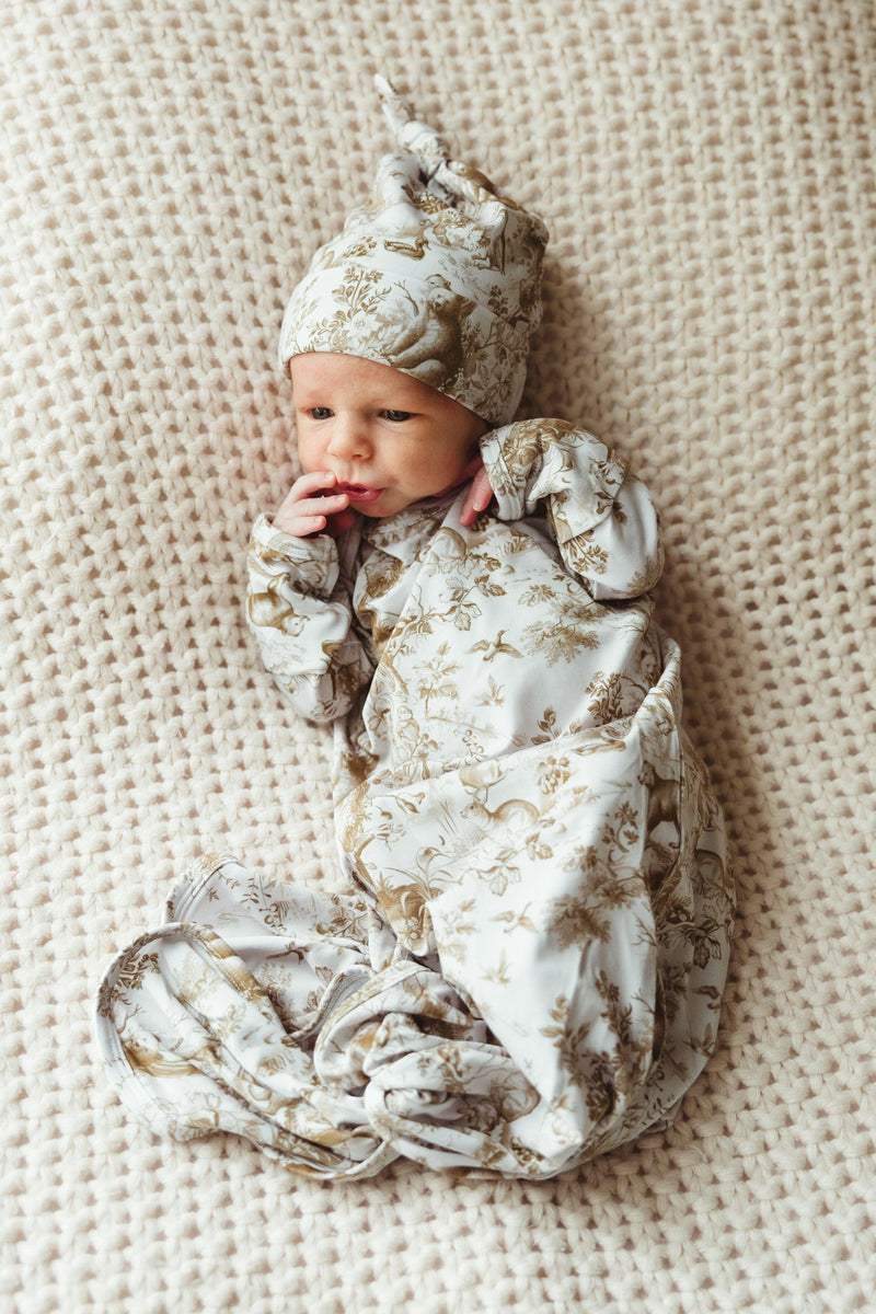 Newborn baby wearing Woodland Toile bamboo knotted gown and matching hat, lying on a textured knit blanket — features soft brown and white woodland animal print with bears.
