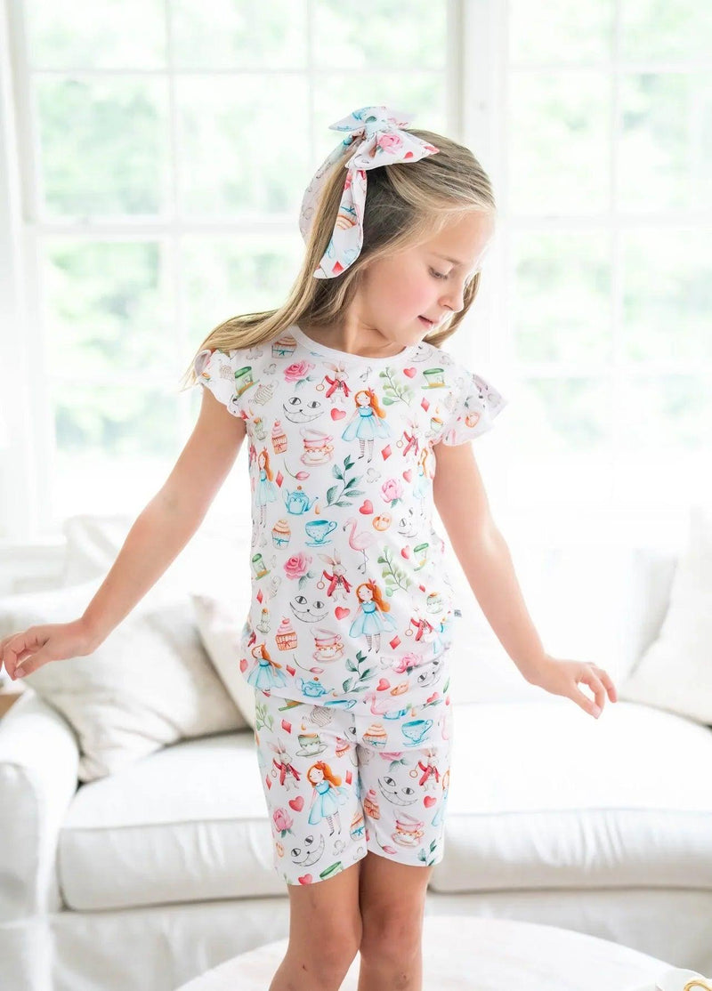 Girl modeling Alice in Wonderland-inspired two-piece pajamas with teacup and cupcake print, shown standing front-facing.