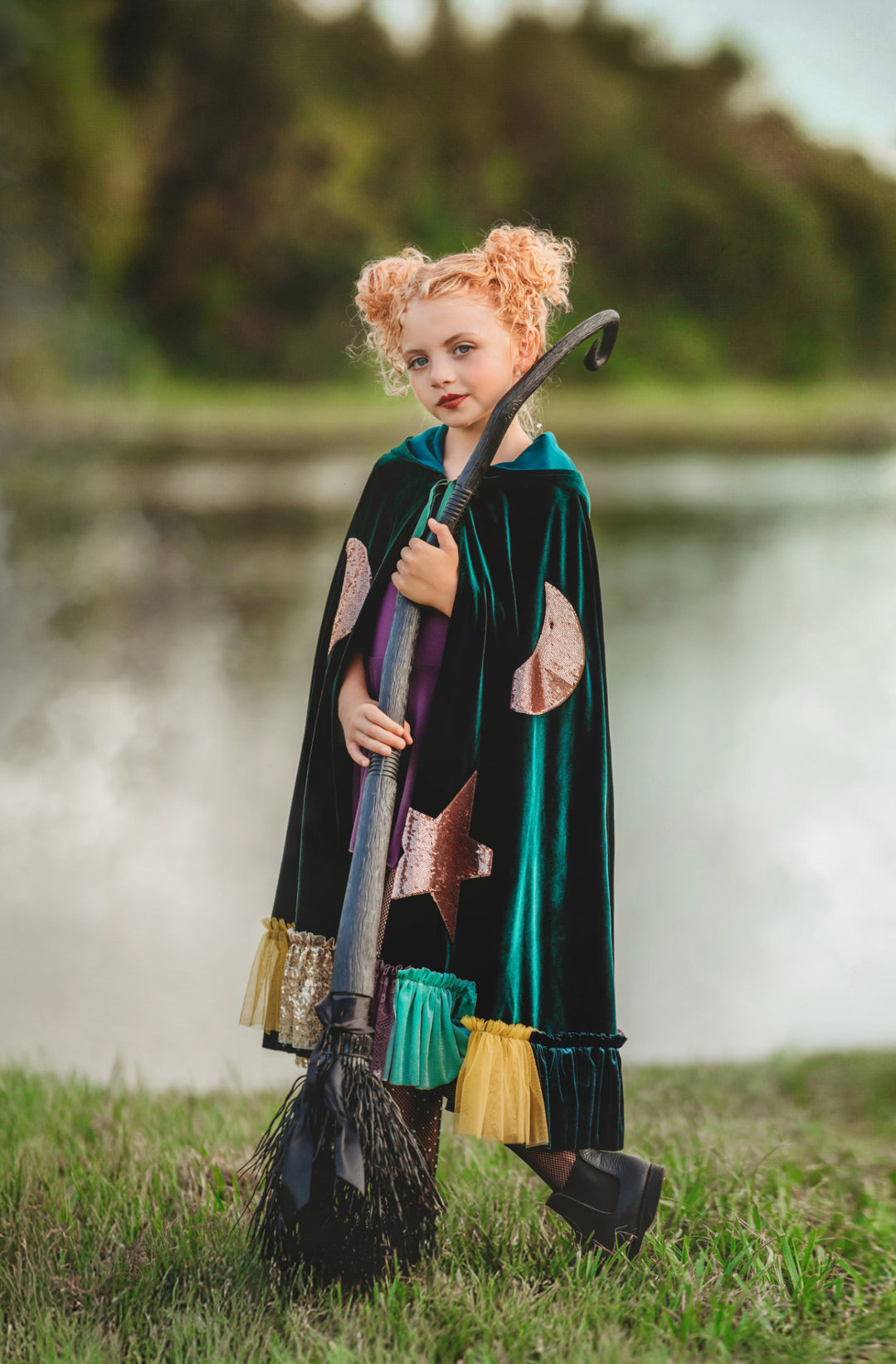 Child in a green  hocus pocus sanderson sisters winifred cape with pink patterns holding a broomstick outdoors by a lake.