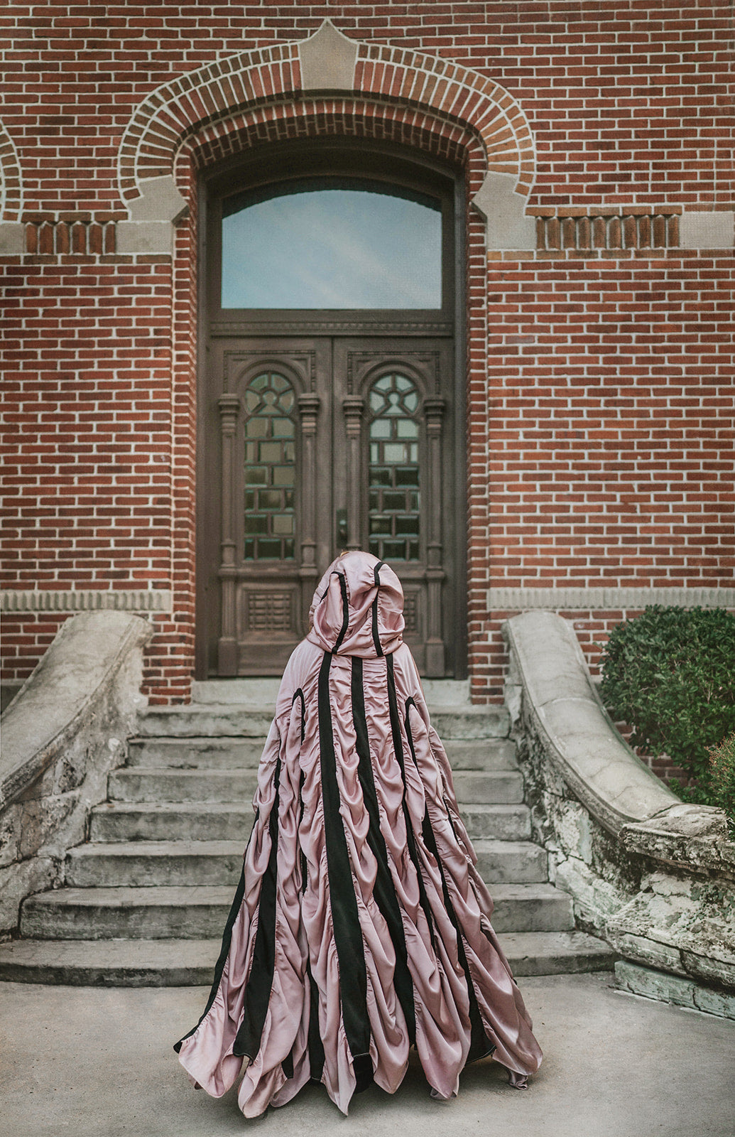 A person viewed from behind wearing a voluminous, floor-length draped vampire coffin cape for halloween in dusty pink and black, standing on stone steps in front of a red brick building with a dark wooden door.