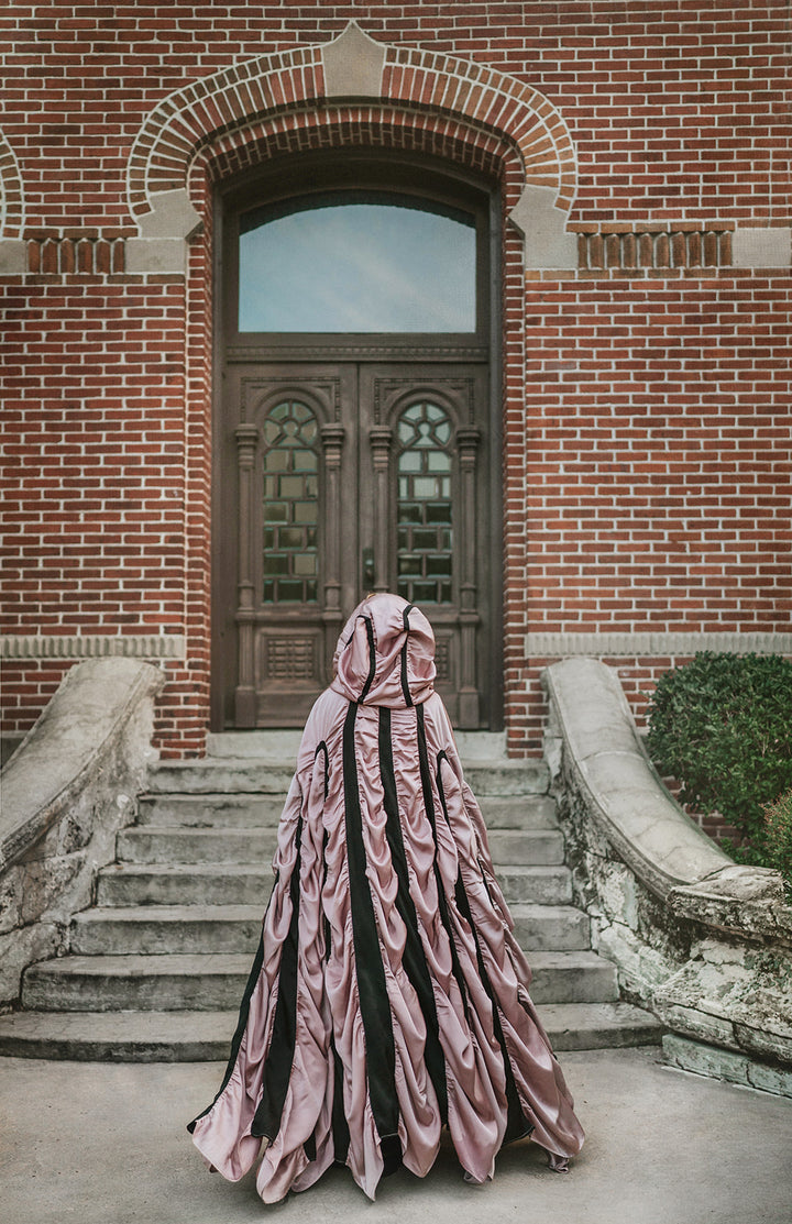 A person viewed from behind wearing a voluminous, floor-length draped vampire coffin cape for halloween in dusty pink and black, standing on stone steps in front of a red brick building with a dark wooden door.
