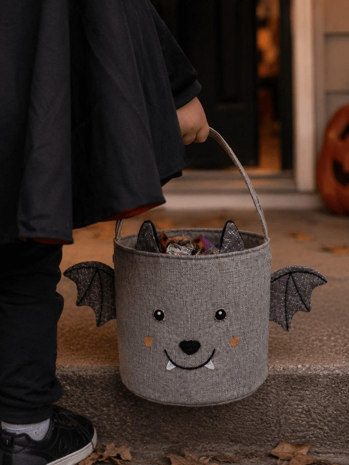 A child wearing a black costume holds a gray felt bat-shaped  trick or treat candy bucket with black bat wings, a stitched face, and a handle, standing on concrete steps in front of a doorway.