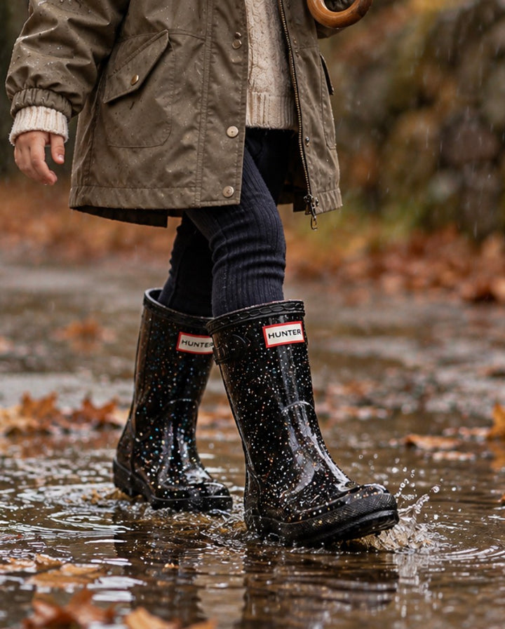 Child wearing Hunter Boots Kids First black glitter waterproof rain boots while walking through a rainy autumn puddle