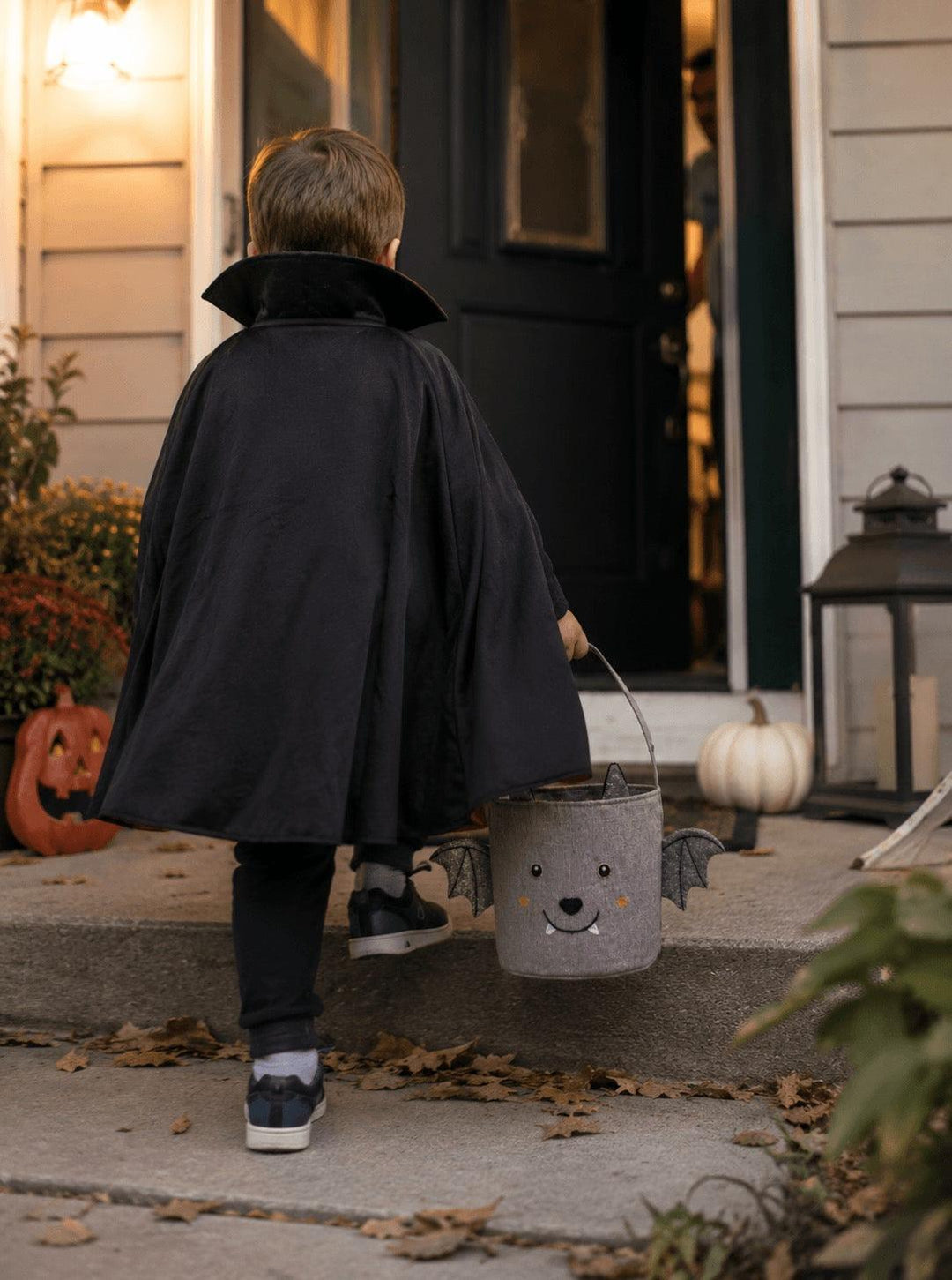 A child viewed from behind wearing a black vampire cape and walking up front steps toward a house door, while holding a gray bucket with a bat face and small wings.