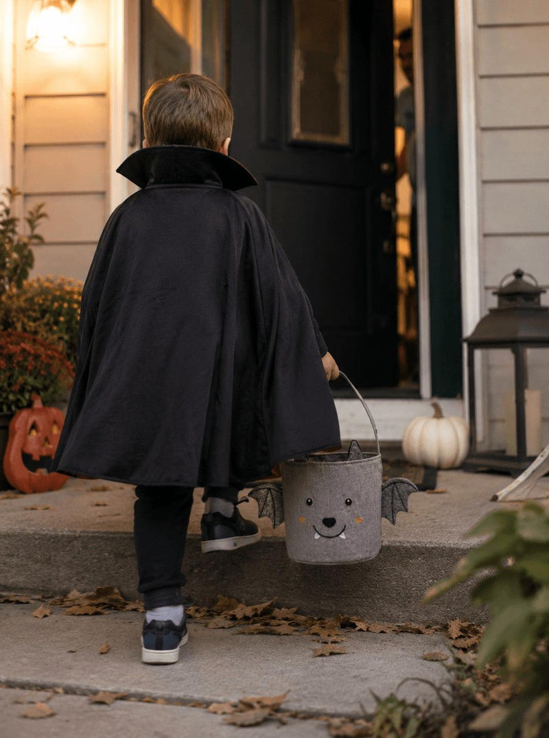 A child viewed from behind wearing a black vampire cape and walking up front steps toward a house door, while holding a gray bucket with a bat face and small wings.