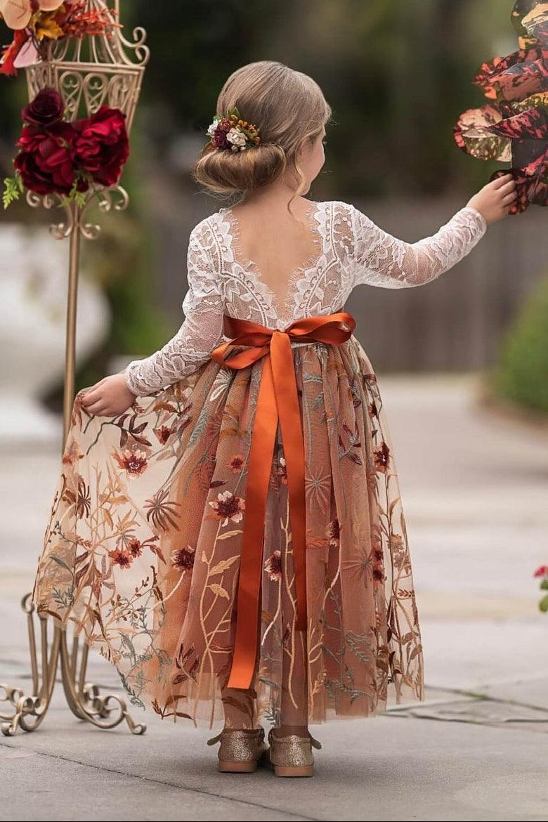 A young girl stands with her back to the camera in a garden, wearing a long-sleeve lace floral  flower girl dress with a sheer tulle skirt, a large orange satin bow at the waist, and brown shoes. She is reaching toward a large floral arrangement on a white planter.