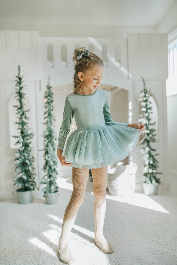 Young girl in a sage green tutu dress standing in a room with decorative plants.