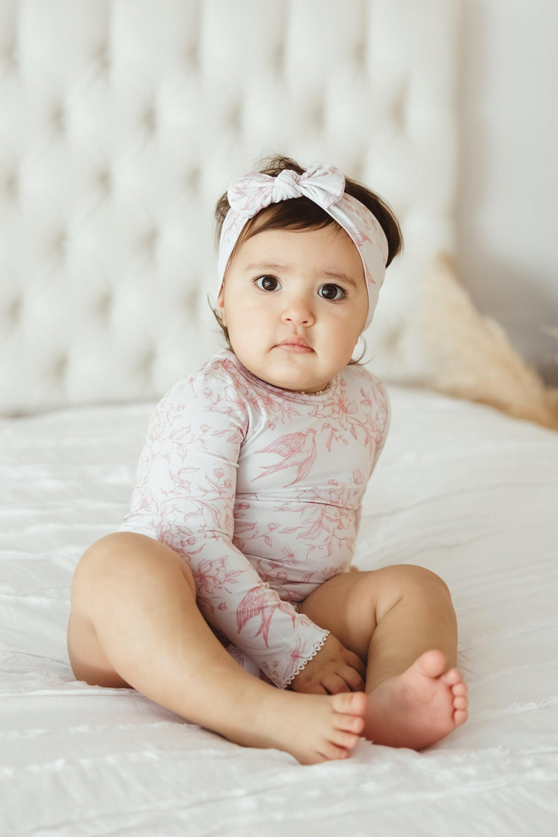 Little girl sitting in Botanical Lullaby long sleeve bamboo baby bodysuit, featuring a delicate off-white and rose toile print with birds, flowers, and bees — soft, breathable, and perfect for everyday wear or gifting.
