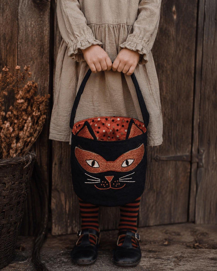 A child wearing a beige dress and striped socks holds a black cat-shaped fabric trick or treat bucket bag with a red polka-dot interior, set against a rustic wooden door background.