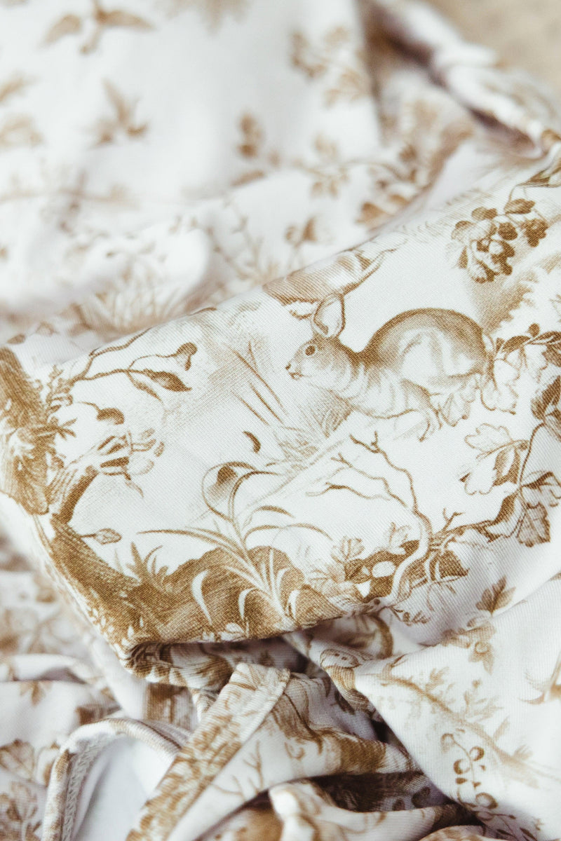 Close-up of Woodland Toile print on bamboo newborn gown, showing detailed forest animals including bears and foliage in soft brown tones on ivory fabric.
