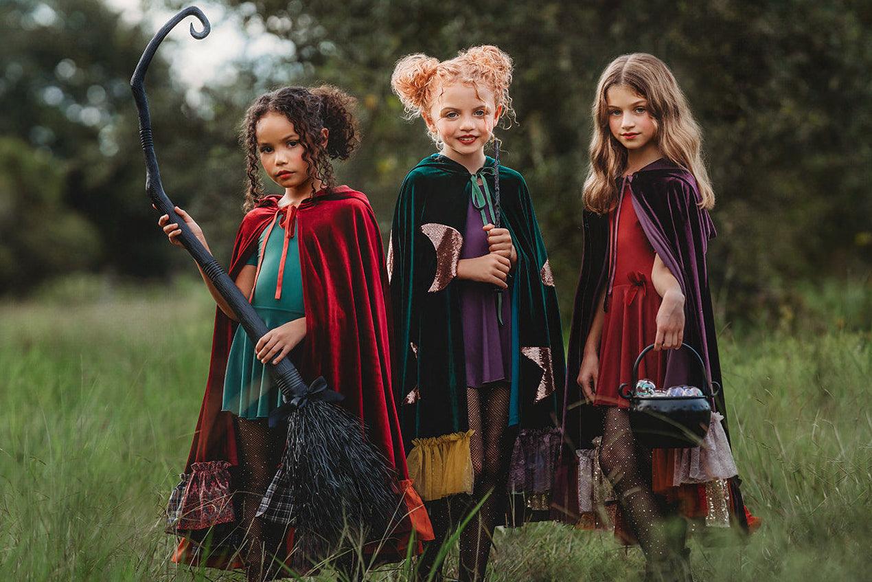 Three young girls dressed as hocu pocus sisters witches stand in a grassy field with trees in the background. The girl on the left, Mary wears a red cape and holds a black bow, the middle girl , Winifred wears a dark green cape, and the girl on the right , Sarah wears a deep purple cape.