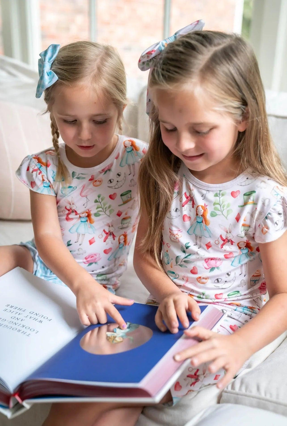 Two little girls reading Alice in Wonderland book while wearing matching Adventures with Alice bamboo pajamas.