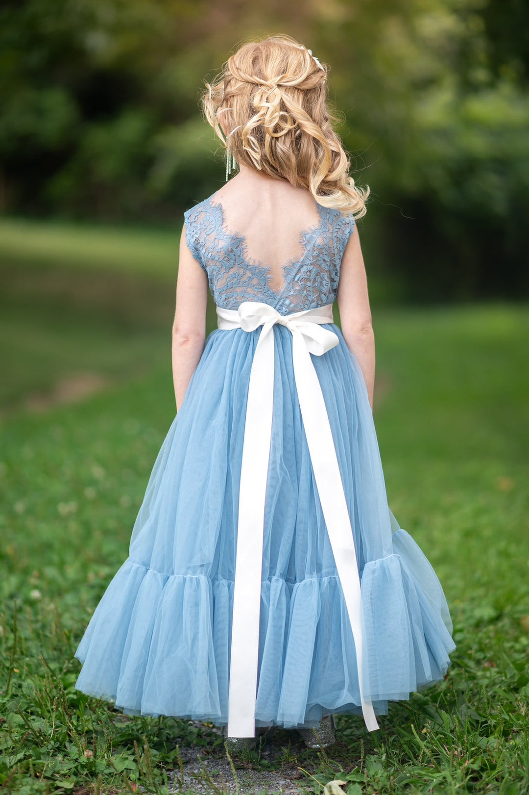 Young girl in a dusty blue flower girl dress with a white ribbon sash standing in a grassy area.