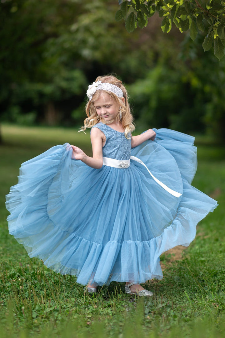 Young girl in a dusty blue flower girl dress with a white belt standing on grass with a blurred green background