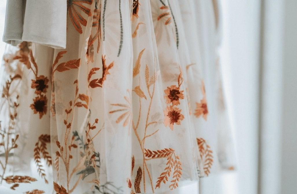 A close-up of a sheer white tulle skirt or drape decorated with embroidered botanical patterns in warm rust orange, tan, and muted green, hanging against a softly blurred light background.