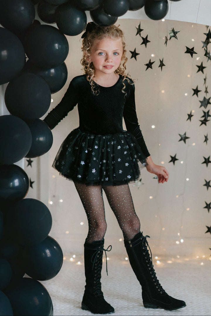 Girl wearing black velvet long sleeve tutu dress with star tulle skirt, standing in front of black balloon arch.