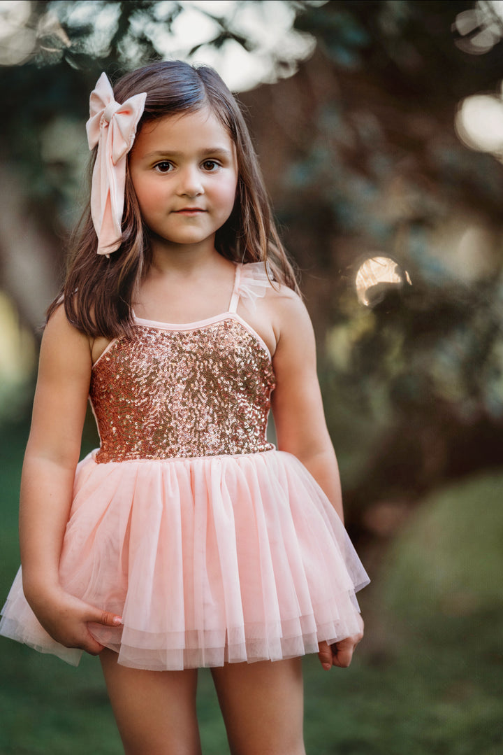 Little girl wearing blush pink sequin tutu dress outdoors, smiling softly.