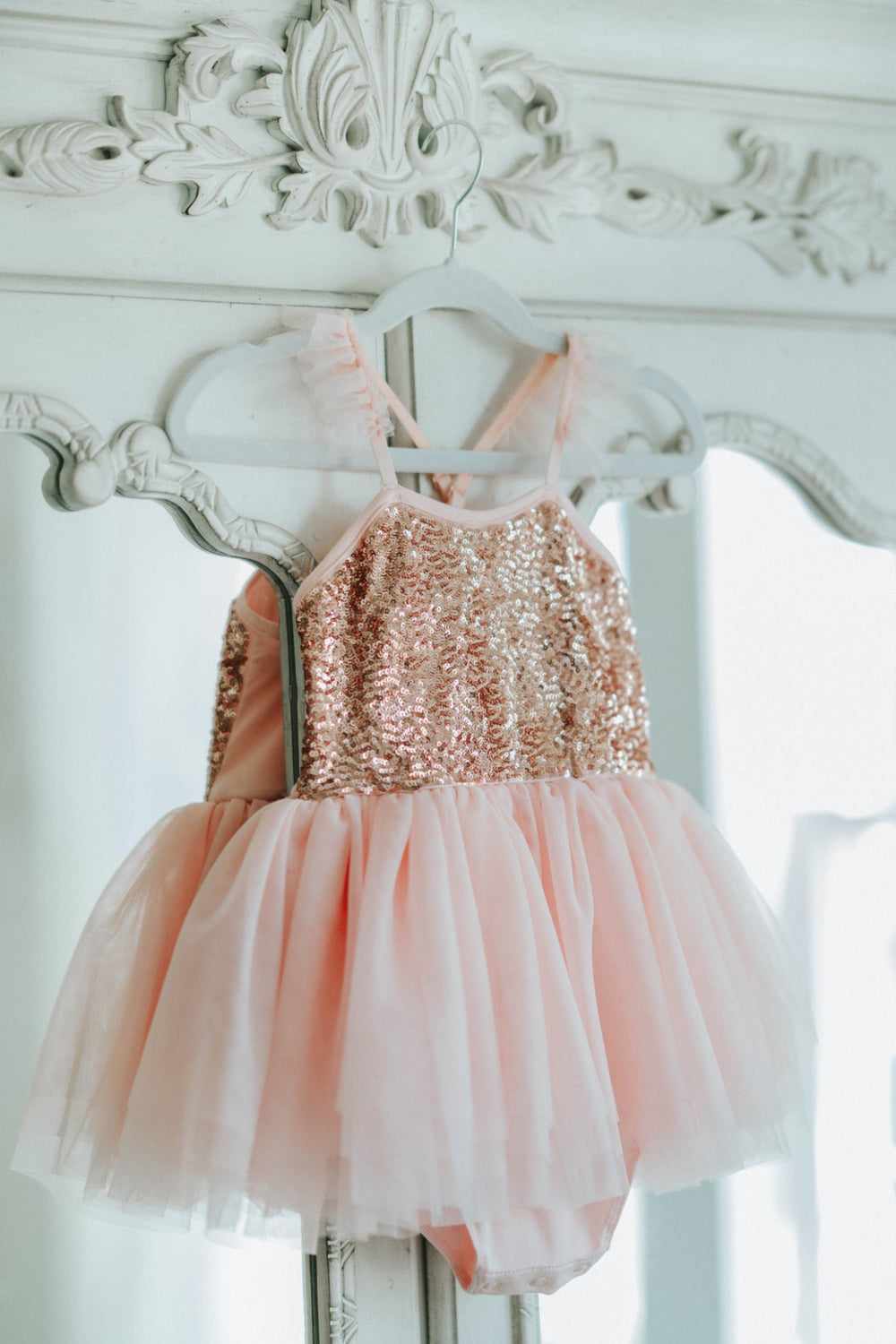 Blush pink sequin bodice tutu dress hanging on antique white wardrobe.
