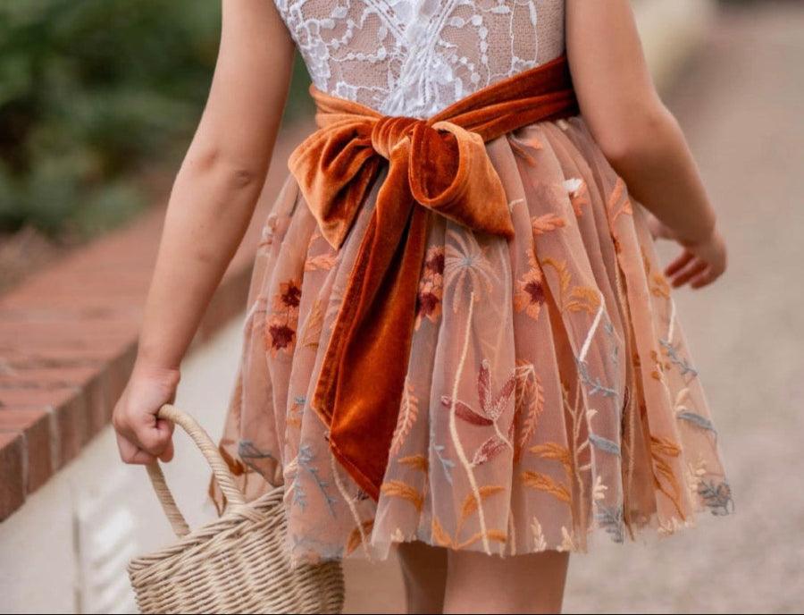 A child model shown from behind wearing a sleeveless white lace V-neck dress with a tan tulle skirt featuring leaf embroidery and a large rust-orange velvet bow at the waist. The model is holding a small woven straw basket bag on a paved outdoor walkway.