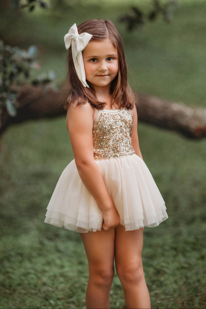 Toddler girl wearing cream and gold sequin tutu dress outdoors, standing and smiling in soft natural light