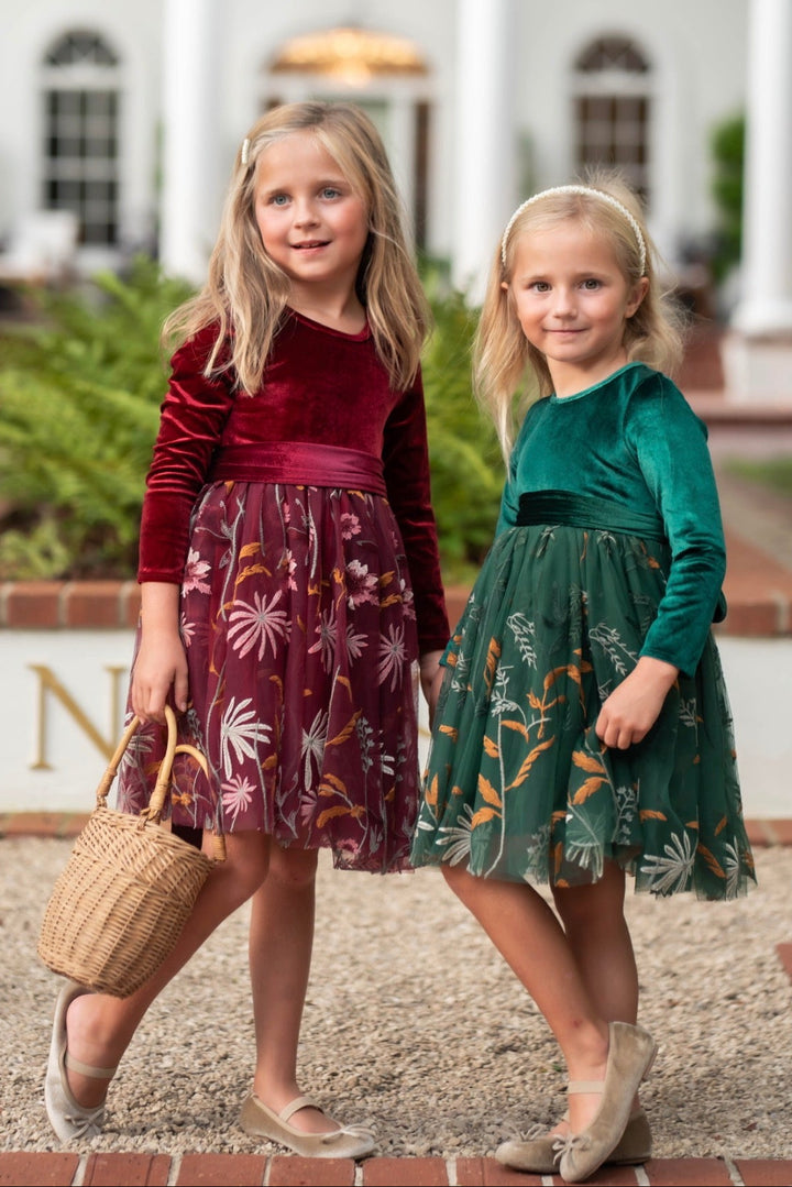 Two young girls wearing elegant velvet and tulle dresses standing in front of a grand white building. The girl on the left wears a deep burgundy velvet long-sleeve dress with a floral tulle skirt and holds a woven basket.
