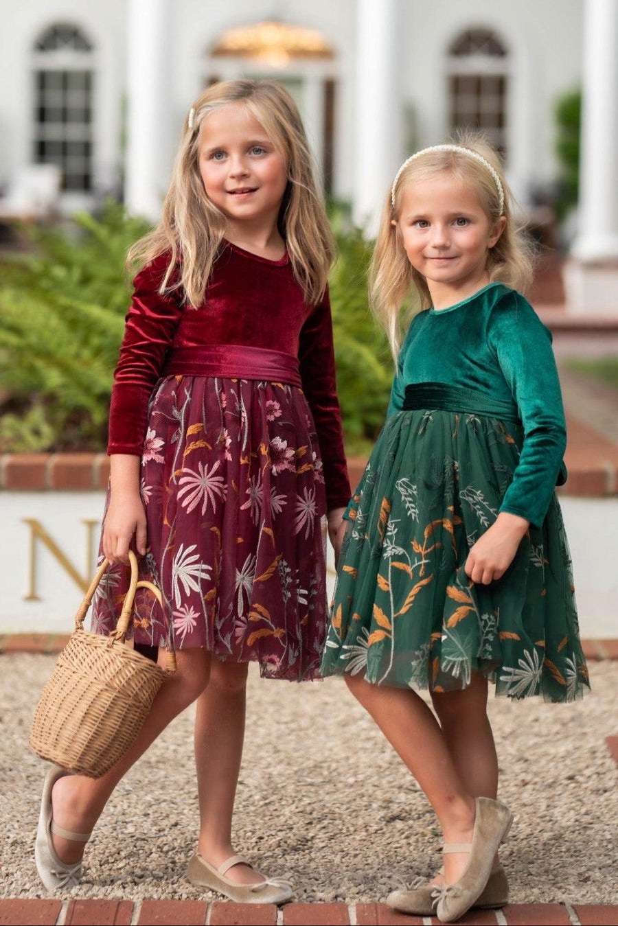 Two young girls wearing elegant velvet and tulle dresses standing in front of a grand white building. The girl on the left wears a deep burgundy velvet long-sleeve dress with a floral tulle skirt and holds a woven basket.