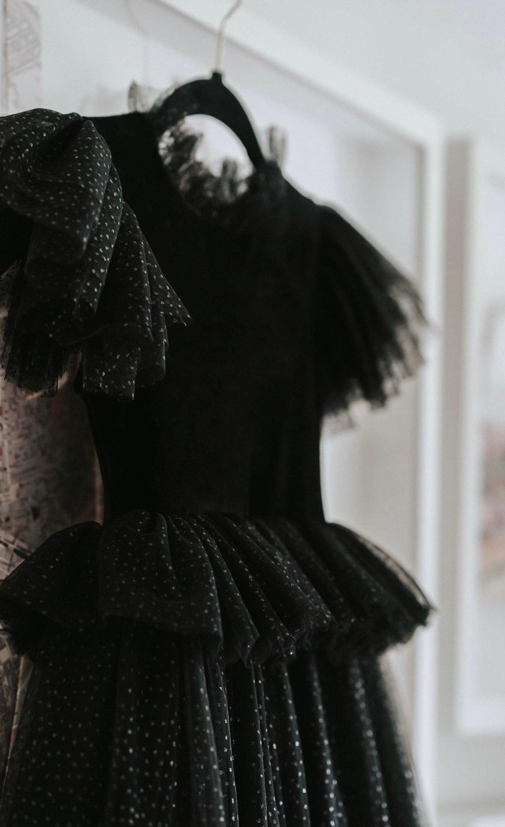 A black ruffled tulle dress with a polka-dot pattern hanging on a black hanger against a white wall.