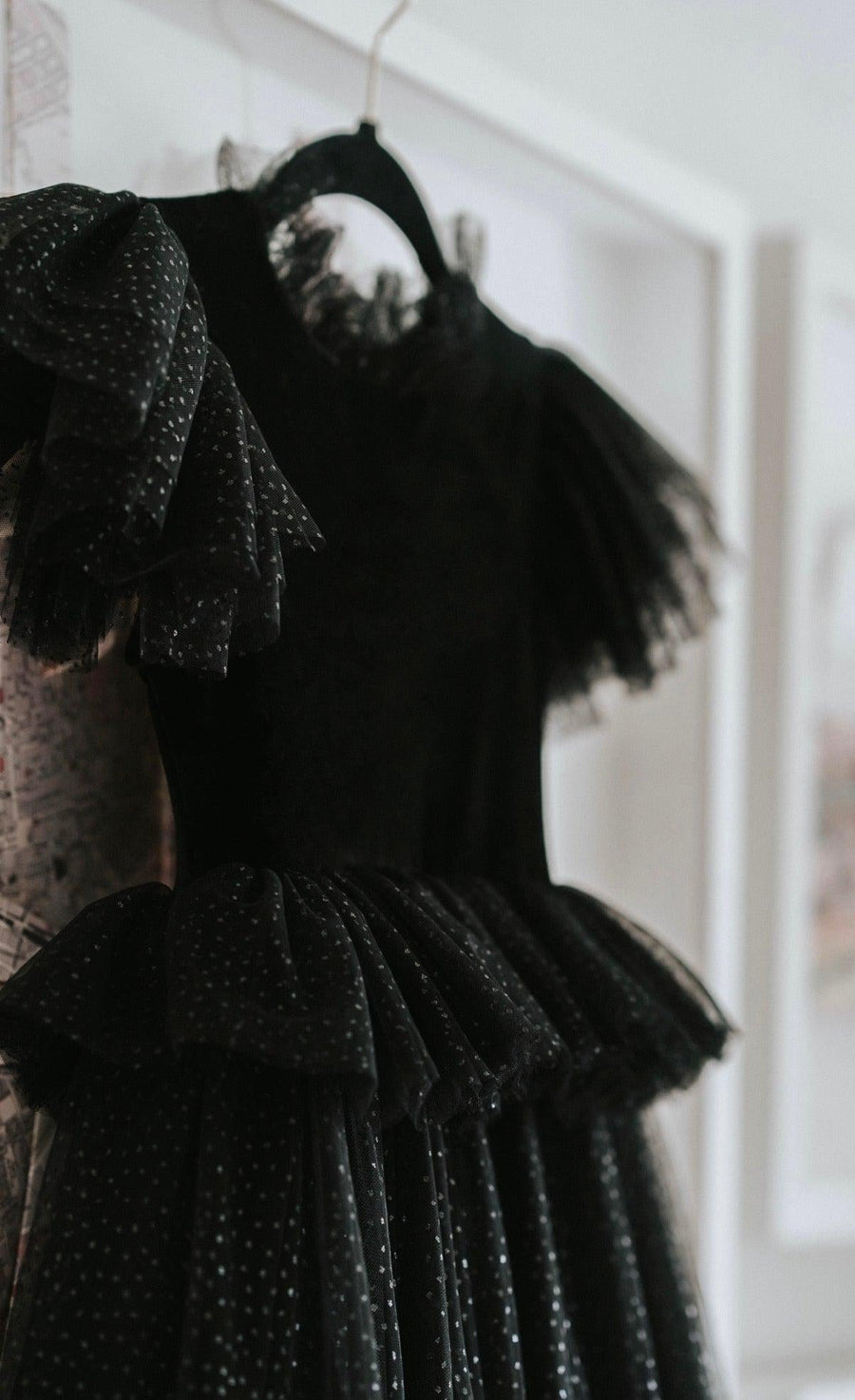 A black ruffled tulle dress with a polka-dot pattern hanging on a black hanger against a white wall.