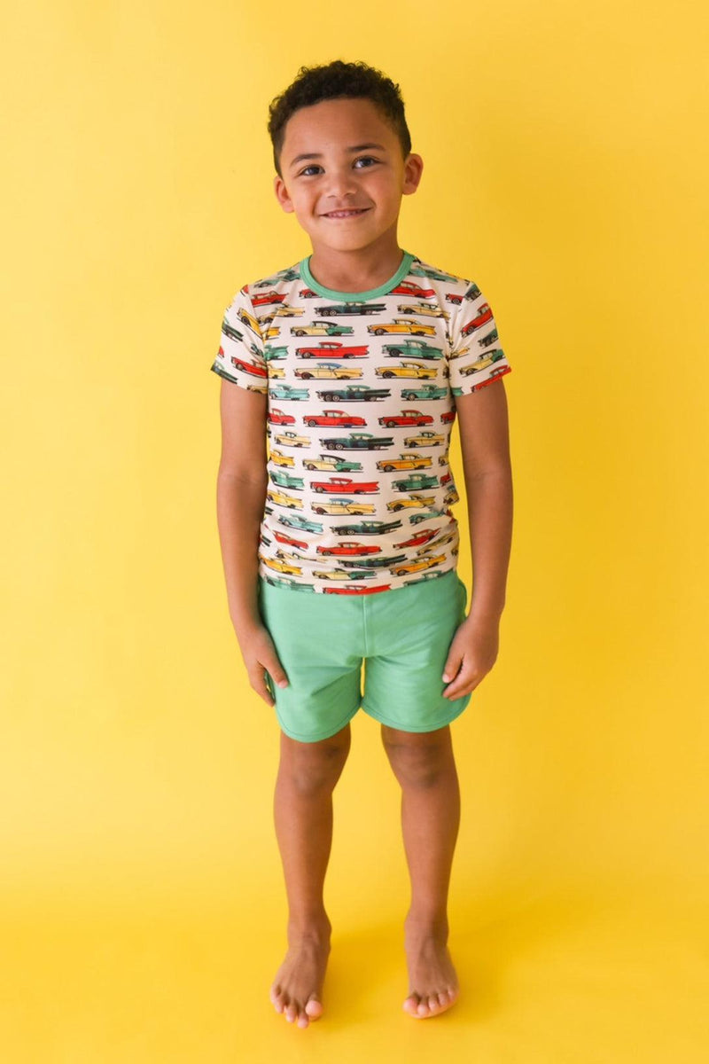Little boy wearing classic cars short sleeve graphic t-shirt and lagoon green French terry shorts, standing full length and smiling in playful summer outfit.
