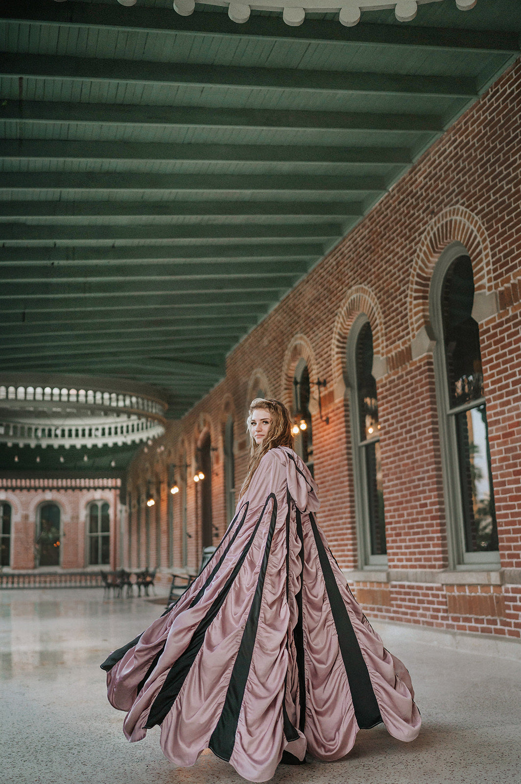 A woman wearing a dramatic floor-length coffin cape costume with pleated dusty mauve and black fabric stands in a large covered brick hall with arched windows and a green ceiling.