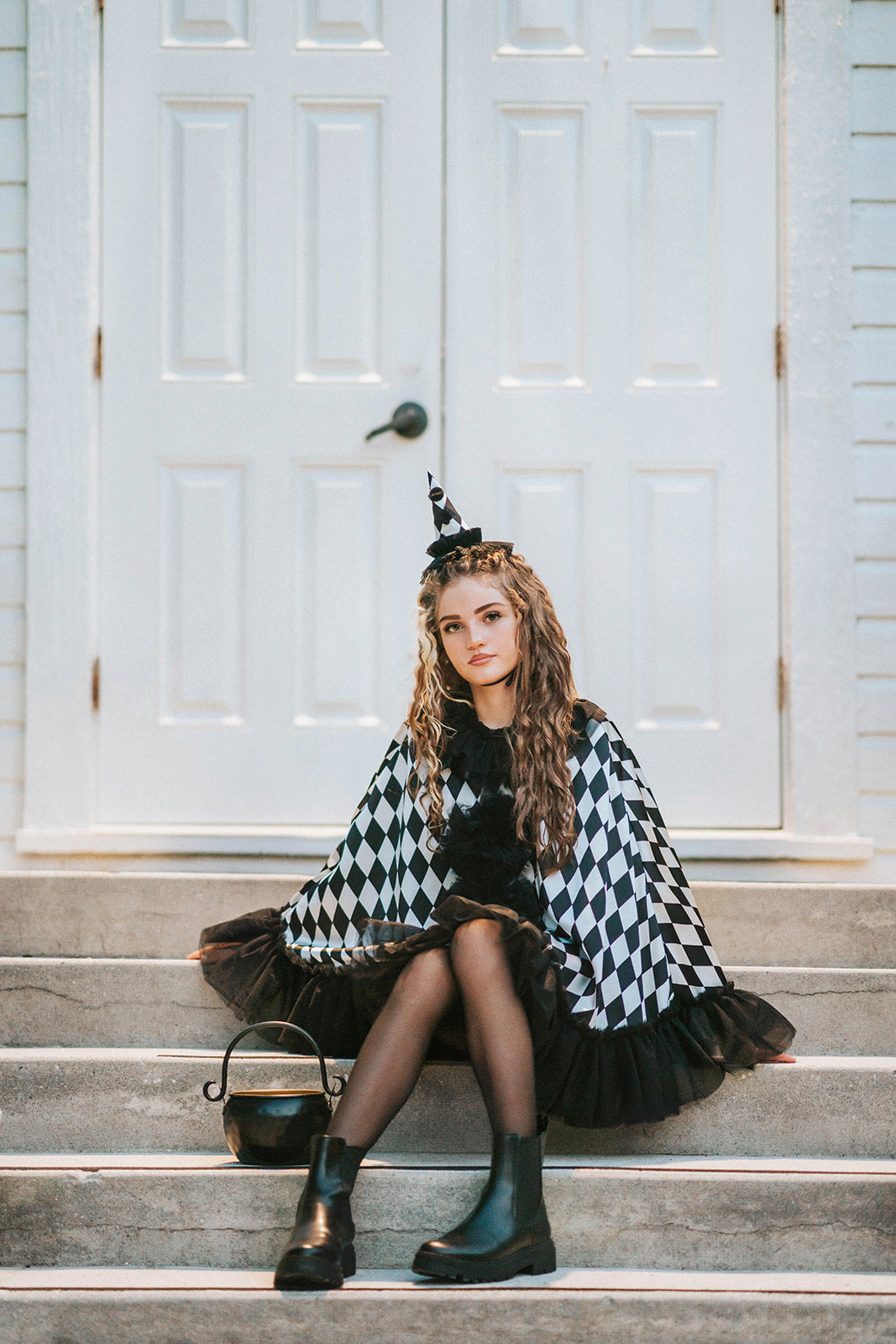 Teen sitting on stone steps wearing the Pirouette black and white harlequin clown cape with black tulle ruffles and matching pointed hat.