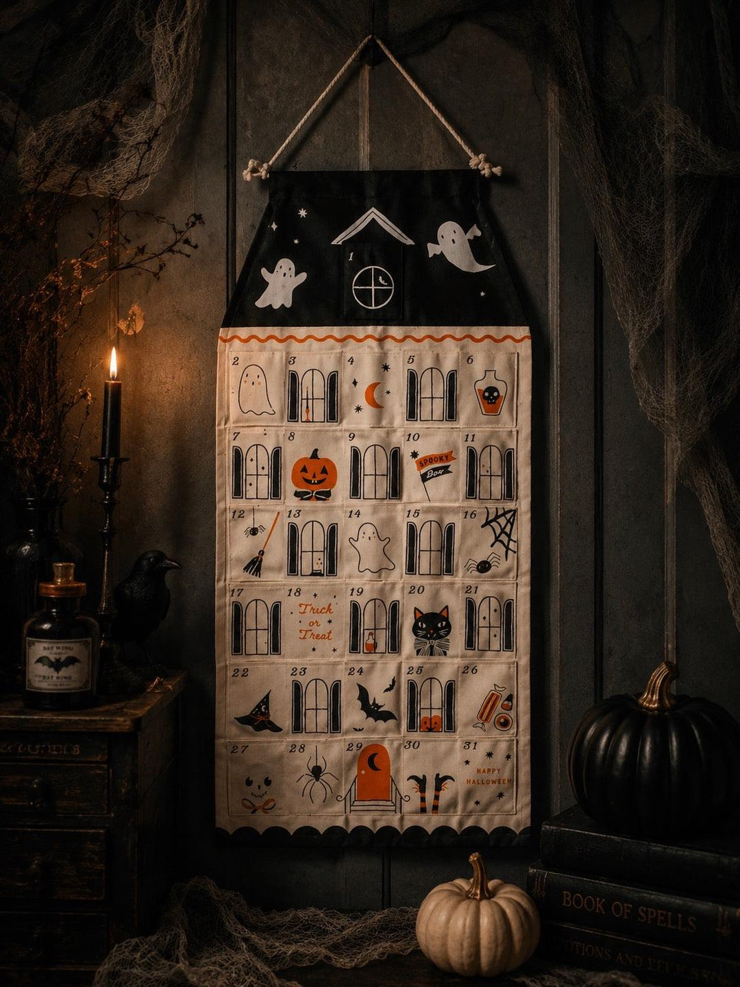 A Halloween-themed fabric wall calendar or advent-style hanging cloth with numbered pockets, ghosts, bats, pumpkins, spiders, and spooky illustrations, displayed against a dark rustic wall with candles, cobwebs, and pumpkins around it.
