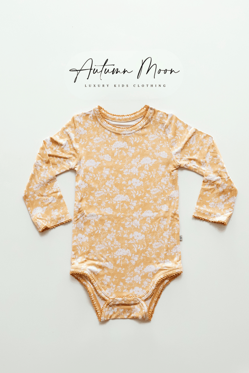 Flat lay of Golden Afternoon bamboo long sleeve baby bodysuit with snap bottom, featuring a golden yellow background with delicate white floral print and scalloped trim — made from buttery soft bamboo viscose, perfect for baby girls and gift registries.
