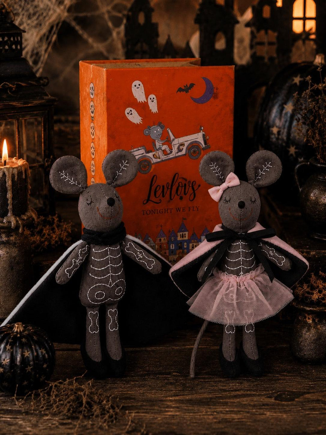 Two Halloween-themed plush mouse dolls dressed as skeletons, standing in front of an orange illustrated box labeled 'Levi's Zombie Witch'. The scene is styled with candles, cobwebs, and dark decorative props on a rustic wooden surface.