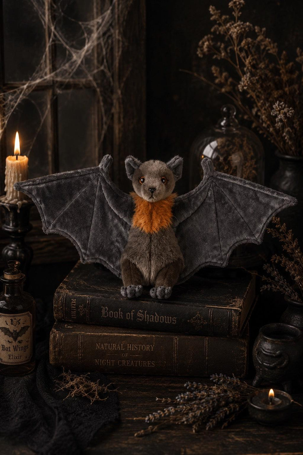 A plush toy bat with dark gray wings and a bright orange collar sits on stacked antique-style books, surrounded by candles, dried flowers, cobwebs, and dark decorative props in a moody Halloween-style scene.