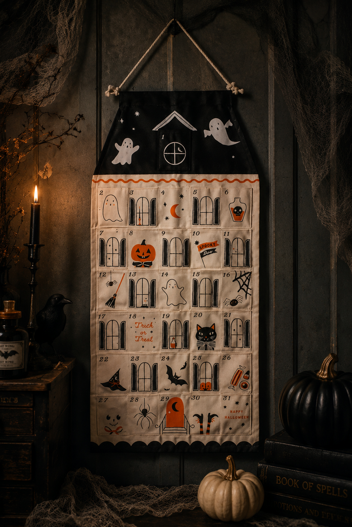 A Halloween-themed fabric wall calendar or advent-style hanging cloth with numbered pockets, ghosts, bats, pumpkins, spiders, and spooky illustrations, displayed against a dark rustic wall with candles, cobwebs, and pumpkins around it.