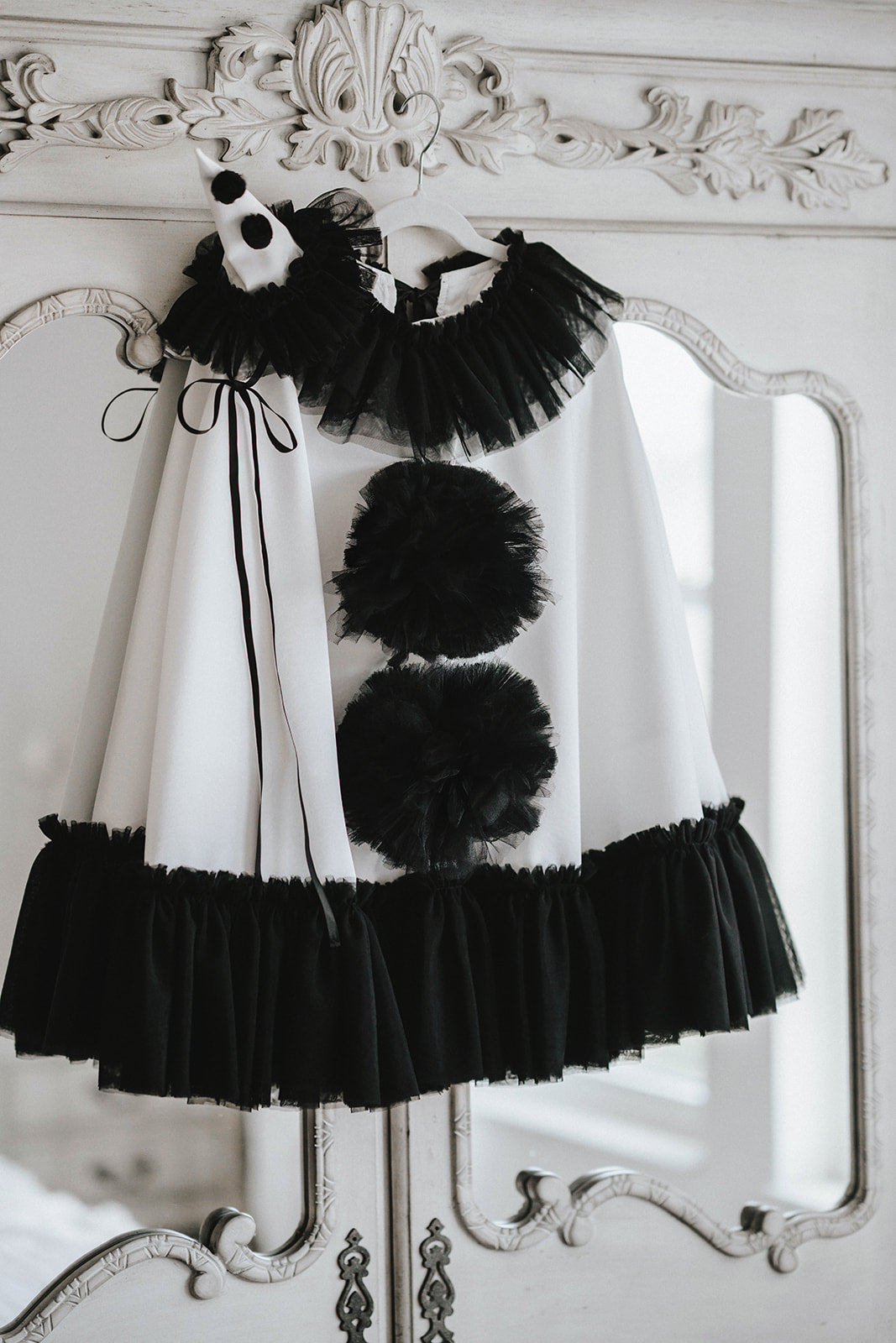 White satin Pirouette clown cape hanging on a closet with black tulle ruffle trim, oversized pom-poms and matching pointed hat.